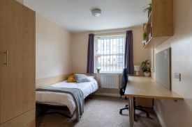 Bedroom containing single bed, desk and chair, wall-mounted pinboard, shelves above the desk, a cupboard, and a wall-mounted radiator